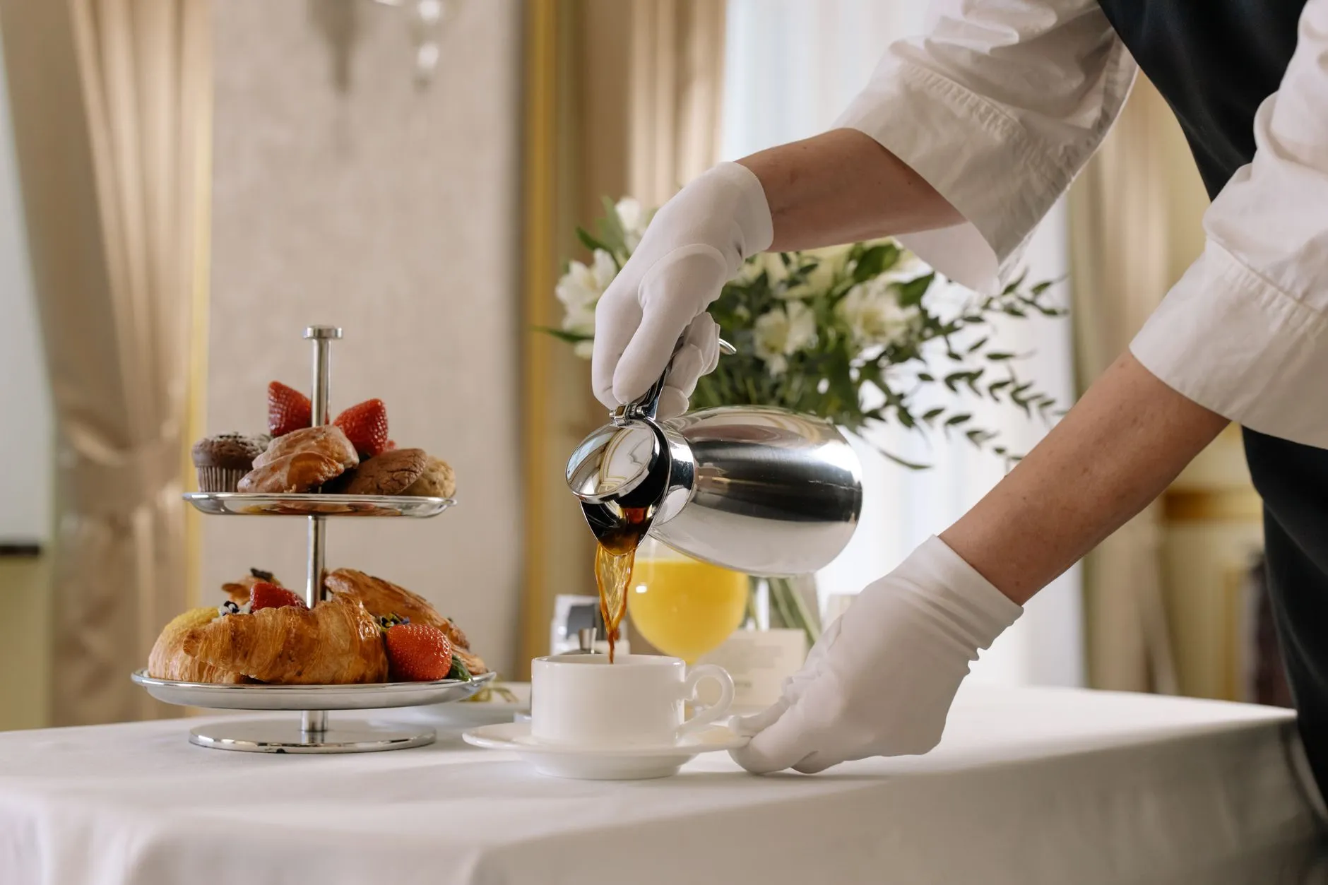Mãos de luvas brancas servindo café numa mesa de café da manhã de hotel, ao lado de uma bandeja de croissants